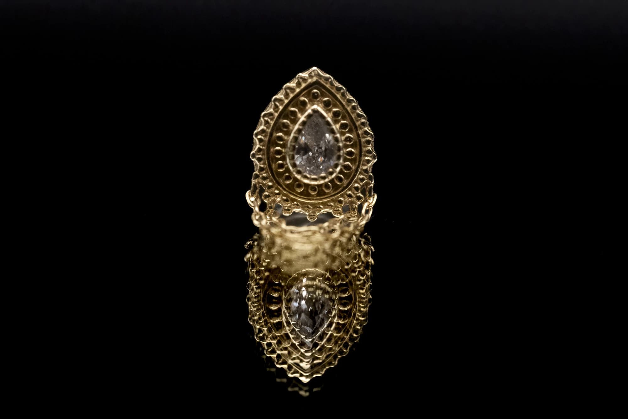 Buddha Jewelry Deity with an ornate pear-shaped gold setting and fine chains reflected on black glass